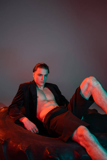 provokatív és divatos fiatalember fekete rövidnadrágban és divatos blézer ing nélküli izmos test ül hatalmas gumiabroncs szürke háttér piros világítás - Fotó, kép