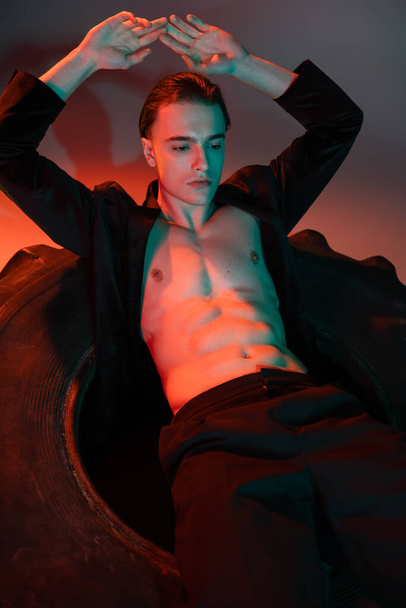 young and sexy man wearing black blazer on shirtless muscular torso, sitting on huge tire and posing with hands above head on grey background with red lighting - Foto, Imagem