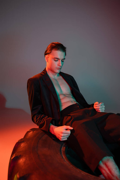 young, good looking and sexy man in black trendy blazer on shirtless muscular torso sitting and posing on huge tire on grey background with red lighting - Фото, изображение