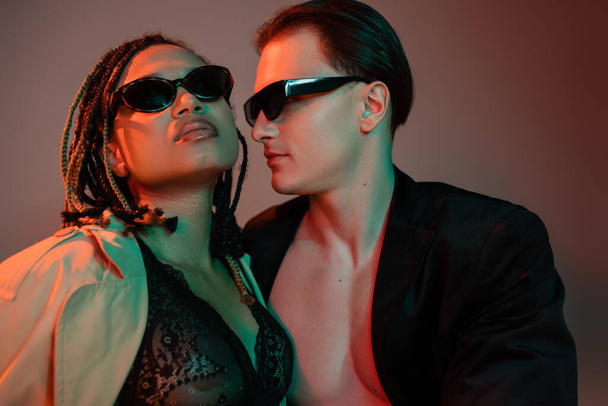 сексуальная межрасовая пара в темных солнцезащитных очках, харизматичный мужчина в черном пиджаке и африканская американка в кружевном белье и бежевом плаще на сером фоне с красным освещением - Фото, изображение