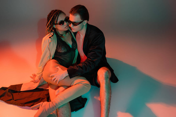 fashionable man in dark sunglasses and black blazer seducing african american woman in lace bodysuit and beige trench coat while hugging her leg on grey background with red lighting - Foto, Bild