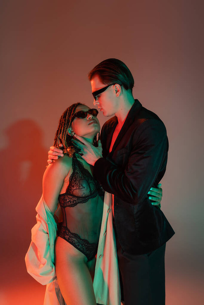 young and confident man in dark sunglasses and black blazer embracing tempting african american woman in lace bodysuit and beige trench coat on grey background with red lighting - Foto, afbeelding