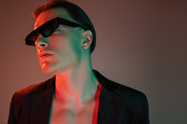 young, self-assured and bare-chested man in black blazer and dark fashionable sunglasses standing and looking away on grey background with red lighting - 写真・画像