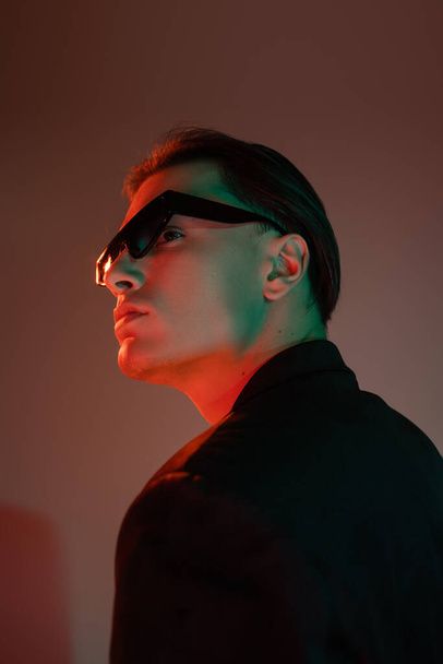 молодой и харизматичный мужчина с брюнетками, позирующий в темных модных солнцезащитных очках и черном блейзере, отворачиваясь на сером фоне с красным освещением - Фото, изображение