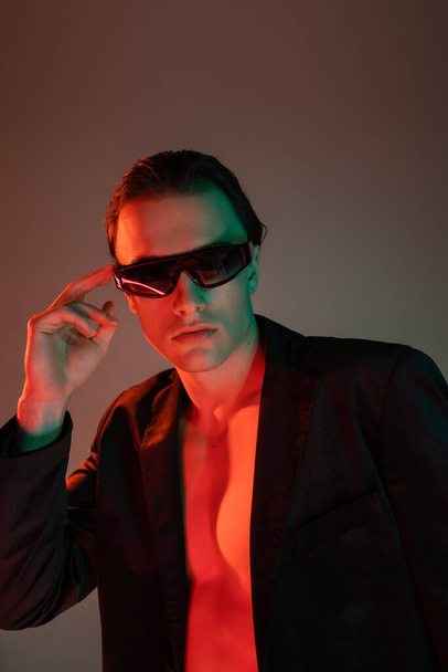 young, bare-chested and appealing man in black blazer adjusting dark fashionable sunglasses and looking at camera on grey background with red lighting - Photo, Image