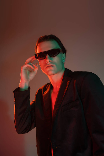 young and good looking man with brunette hair adjusting dark trendy sunglasses while posing in black blazer and looking at camera on grey background with red lighting - Fotografie, Obrázek