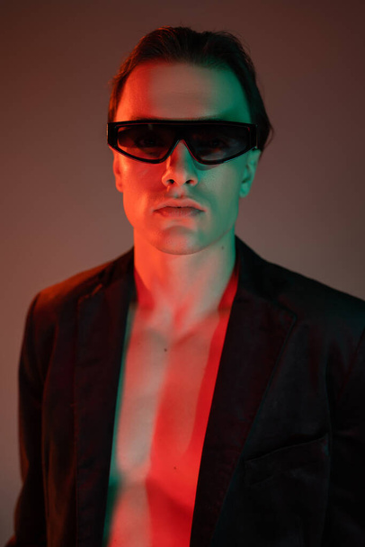 portrait of young and fashionable man in dark sunglasses and stylish blazer looking at camera while standing and posing on grey background with red lighting - Foto, Imagen