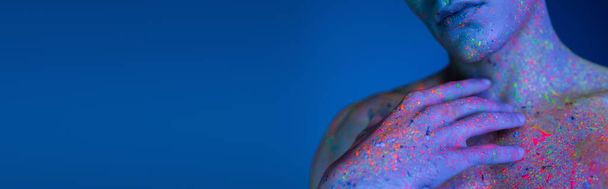 vista parcial del hombre joven sosteniendo la mano cerca del pecho desnudo mientras posa en pintura corporal de neón radiante y multicolor sobre fondo azul con efecto de iluminación cian, pancarta - Foto, Imagen