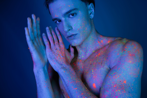 youthful, self-assured and shirtless man in vibrant and colorful neon body paint holding hands near face while looking at camera on blue background with cyan lighting effect - Fotografie, Obrázek
