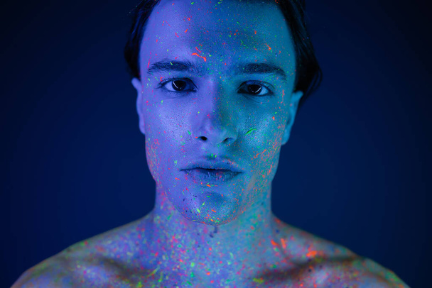 портрет уверенного, уверенного в себе человека с голыми плечами, позирующего в яркой и красочной неоновой краске тела, глядя на камеру на синем фоне с эффектом синего освещения - Фото, изображение