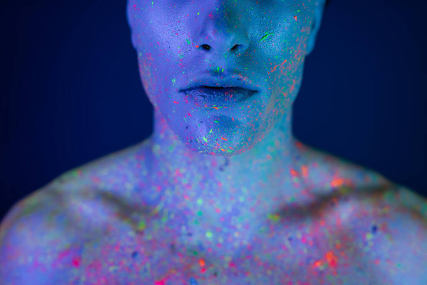 partial view of young and shirtless man in multicolored and vibrant neon body paint standing and posing on blurred blue background with cyan lighting effect - Foto, Imagem