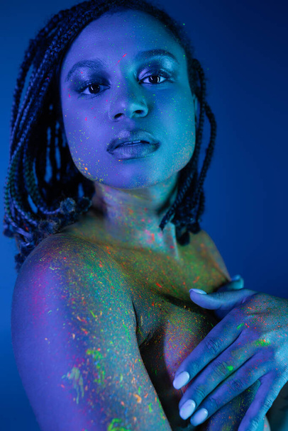 expressive and charming african american woman in radiant and multicolored neon body paint covering breast with hands and looking at camera on blue background with cyan lighting effect - Photo, Image