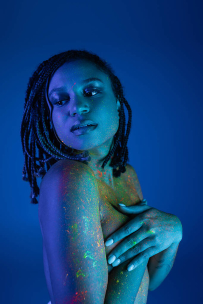 young and tempting bare-chested african american woman with dreadlocks, in colorful neon body paint, covering breast with hands and looking away on blue background with cyan lighting effect - Photo, Image