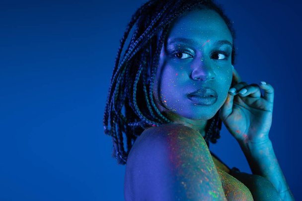 giovane e attraente donna afro-americana in vernice multicolore al neon corpo tenere la mano vicino al viso, mentre guardando lontano su sfondo blu con effetto luce ciano - Foto, immagini