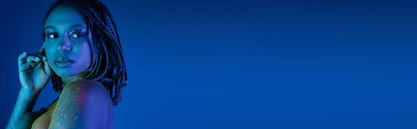 sensuale donna afroamericana con dreadlocks e spalla nuda, in vernice colorata al neon raggiante che distoglie lo sguardo su sfondo blu con effetto ciano, banner - Foto, immagini