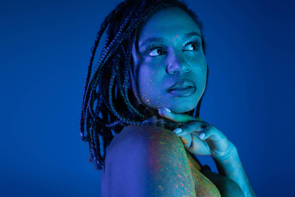 Büyüleyici ve seksi Afro-Amerikalı kadın portresi. Rasta desenli, neon renkli vücut boyası içinde. Mavi arka planda çenenin yanında el ele tutuşurken yüzünü çeviriyor. - Fotoğraf, Görsel