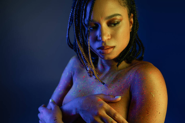 aantrekkelijk Afrikaans amerikaanse vrouw met dreadlocks bedekken borst met handen terwijl poseren in stralende en kleurrijke neon body paint op blauwe achtergrond met gele lichteffect - Foto, afbeelding