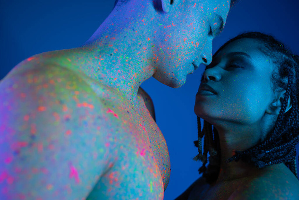 low angle view of young and sensual interracial couple with bare shoulders, in colorful neon body paint standing face to face with closed eyes on blue background with cyan lighting - Foto, Imagem