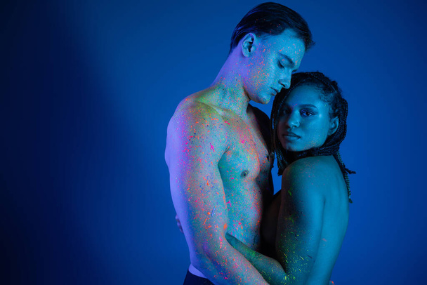 youthful african american woman with dreadlocks, in colorful neon body paint, embracing sexy shirtless man with muscular body and looking at camera on blue background with cyan lighting - Photo, Image