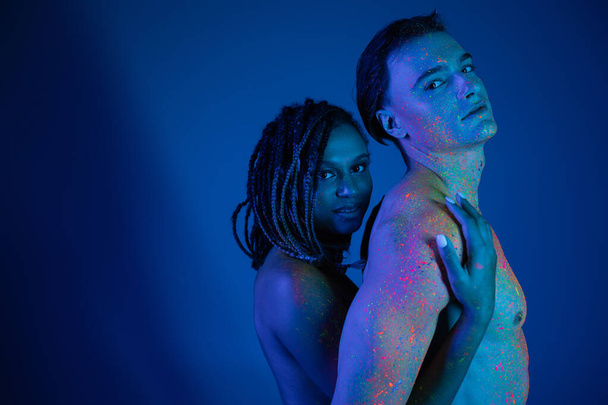 junges multikulturelles Paar in bunter Neon-Körperfarbe mit Blick in die Kamera auf blauem Hintergrund mit Cyanbeleuchtung, afrikanisch-amerikanische Frau umarmt hemdlosen Mann mit muskulösem Oberkörper - Foto, Bild