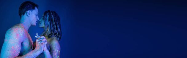 hombre musculoso sin camisa y mujer afroamericana con rastas mirándose el uno al otro y de pie con las manos apretadas en colorida pintura de cuerpo de neón sobre fondo azul con iluminación cian, pancarta - Foto, imagen