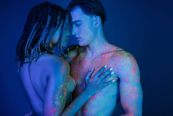 інтимний момент міжрасової пари в барвистій неоновій фарбі тіла, оголена афроамериканська жінка торкається голих грудей без сорочки м'язистого чоловіка на синьому фоні з блакитним освітленням
 - Фото, зображення