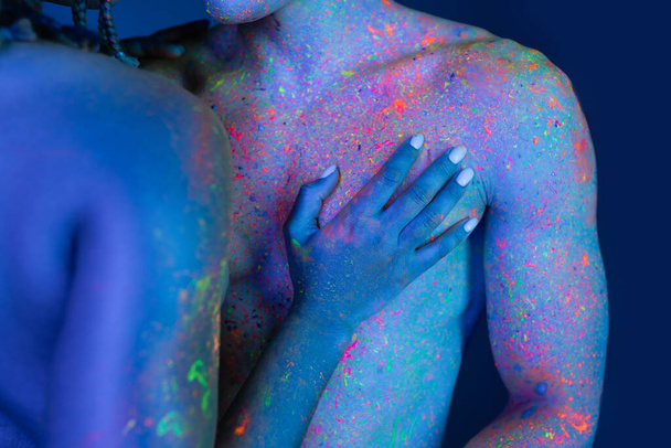 若い情熱的なアフリカ系アメリカ人女性が裸の胸の男に筋肉質の体で触れながらシアン照明と青の背景に立っている部分的なビュー  - 写真・画像