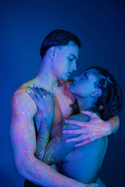youthful interracial couple in colorful neon body paint embracing on blue background with cyan lighting, shirtless man with muscular torso and african american woman with dreadlocks - Photo, Image