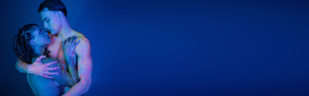 jeugdig en naakt interraciaal paar in kleurrijke neon body paint knuffelen op blauwe achtergrond met cyaan verlichting, intiem moment van charmante Afrikaanse Amerikaanse vrouw en man met gespierd lichaam, banner - Foto, afbeelding