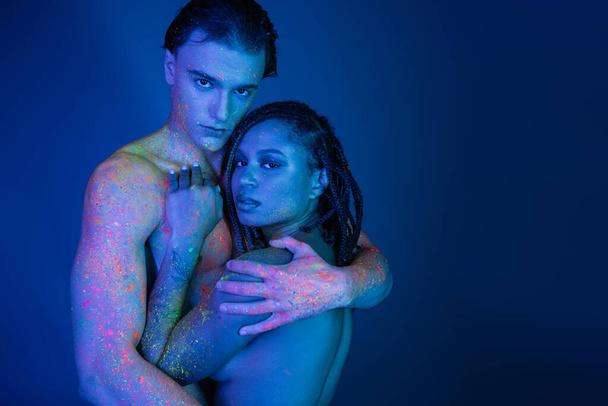 multikulturelles Paar in bunter Neonfarbe, das sich umarmt und in die Kamera blickt auf blauem Hintergrund mit Cyanbeleuchtung, hemdloser Mann mit muskulösem Körper und afrikanisch-amerikanische Frau mit Dreadlocks - Foto, Bild