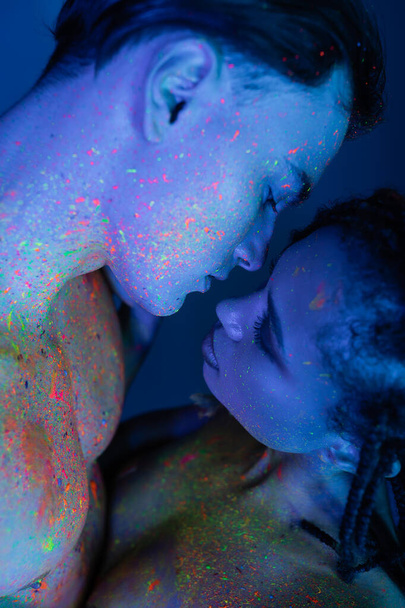 интимный момент молодой мультикультурной пары в цветной неоновой краске тела, обнаженного мужчины и сексуальной африканской женщины на голубом фоне с голубой подсветкой - Фото, изображение