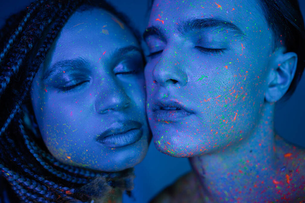 ritratto di giovane coppia multiculturale in colorato neon body paint posa faccia a faccia con gli occhi chiusi su sfondo blu con illuminazione ciano, uomo carismatico e donna afroamericana ipnotizzante - Foto, immagini