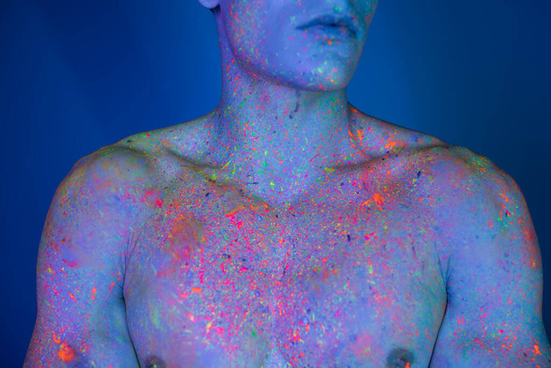 vista parcial del hombre sin camisa, con el pecho desnudo con el cuerpo muscular posando en multicolor y brillante pintura de cuerpo de neón, mientras que de pie sobre fondo azul con efecto de iluminación cian - Foto, imagen