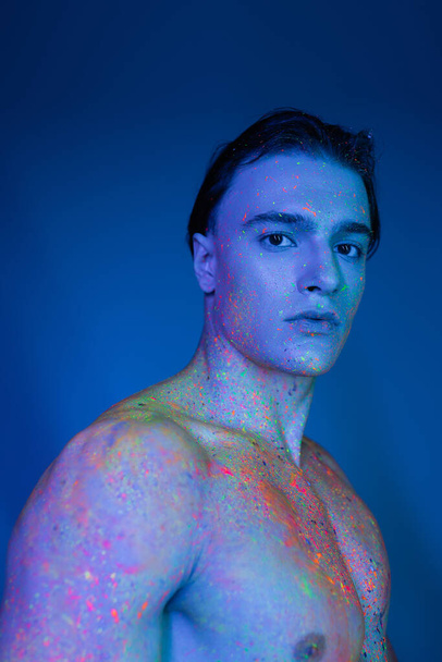 portrait of youthful and handsome shirtless man in multicolored neon body paint looking at camera while standing on blue background with cyan lighting effect - Zdjęcie, obraz