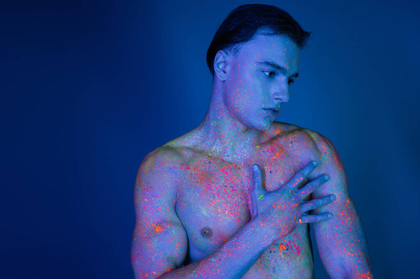 handsome and young shirtless man with muscular torso, in colorful neon body paint, touching bare chest while standing on blue background with cyan lighting effect - Foto, Imagem