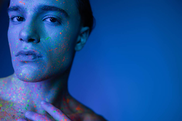 portret van goed uitziende jonge man met zelfverzekerde gezichtsuitdrukking poseren in veelkleurige neon body paint terwijl hij kijkt naar camera op blauwe achtergrond met cyaan lichteffect - Foto, afbeelding