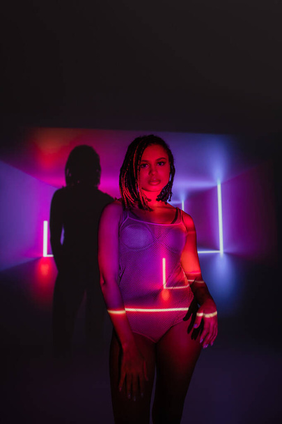 irresistible african american woman in net bodysuit, with stylish dreadlocks looking at camera while standing on abstract black and purple background with neon rays and lighting effects - Photo, Image