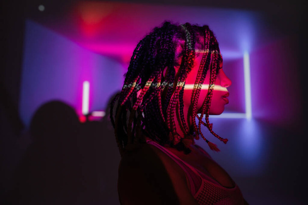 profile of young and charming african american woman with stylish dreadlocks standing on abstract purple background with vibrant neon rays and lighting effects - Foto, afbeelding