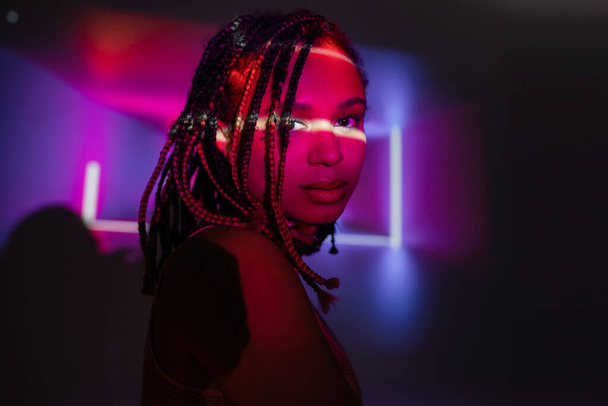portrait of magnetic and appealing african american woman with dreadlocks looking at camera on abstract black and purple background with neon rays and lighting effects - Photo, Image