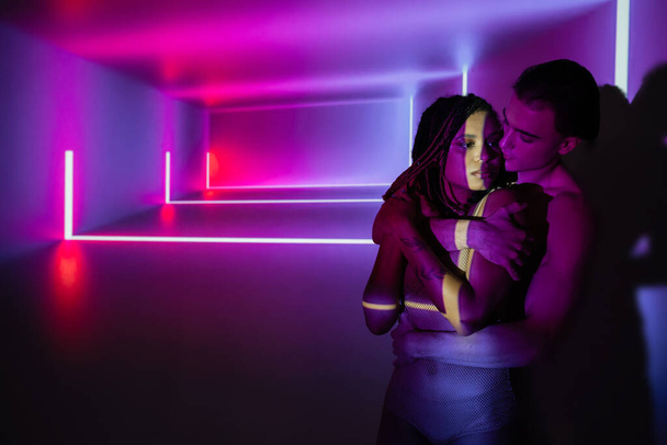 страстная межрасовая пара, харизматичный мужчина и молодая африканская американка с дредами на абстрактном фиолетовом фоне с неоновыми лучами и световыми эффектами - Фото, изображение