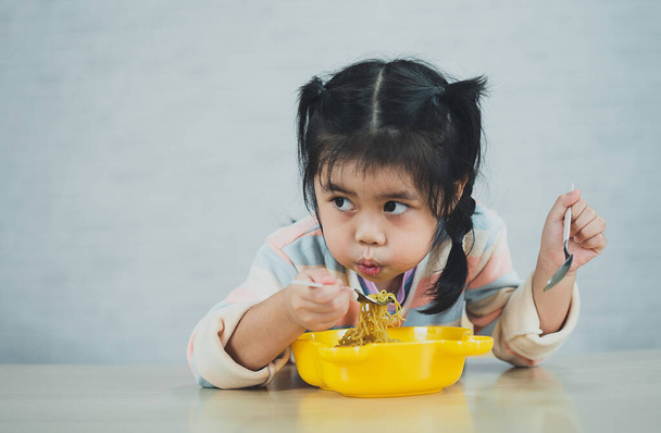 Aasian vauva tyttö nauttia onnellinen käyttäen ruokailuvälineet lusikka ja haarukka syö herkullista nuudeli keittiössä ruokapöydällä. Onnellinen aasialainen vauva tyttö käytännössä syö itse ruokapöydällä. Vauvanruoan käsite - Valokuva, kuva