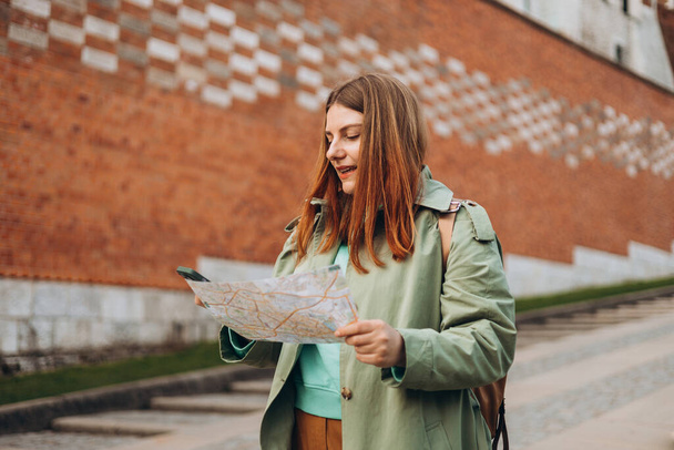 Glückliche rothaarige Frau lächelt zuversichtlich und hält Stadtplan an Straße. Attraktive junge Touristinnen erkunden die neue Stadt. Europa im Frühling bereisen. Banner des urbanen Lebensstils - Foto, Bild
