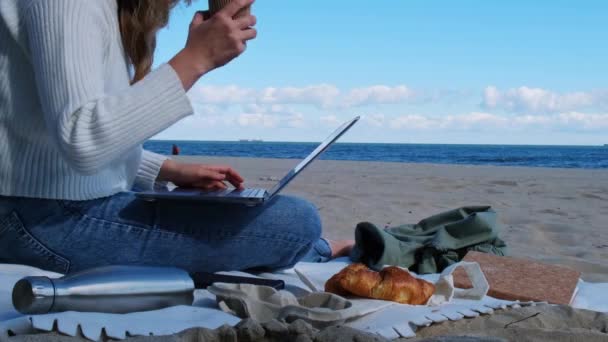 Egy fiatal nő laptopon dolgozik a tengerparton, kilátással a tengerre. Kávézni és croissant-t enni. Student Study munka szabadban meleg őszi tengerparton. Szabadúszó  - Felvétel, videó
