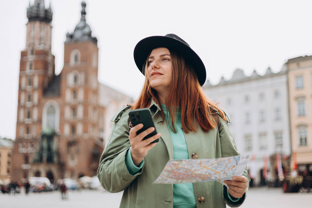 Piękna stylowa kobieta w kapeluszu spacerująca po Rynku w Krakowie w wiosenny dzień i trzymająca telefon komórkowy oraz mapę. Komunikacja telefoniczna. Koncepcja miejskiego stylu życia. Sprawdź portale społecznościowe, rezerwacja hotelu - Zdjęcie, obraz