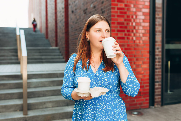 Cheerful young woman wearing blue dress walking outdoors, holding takeaway coffee cup on city background. Urban lifestyle concept. - Φωτογραφία, εικόνα