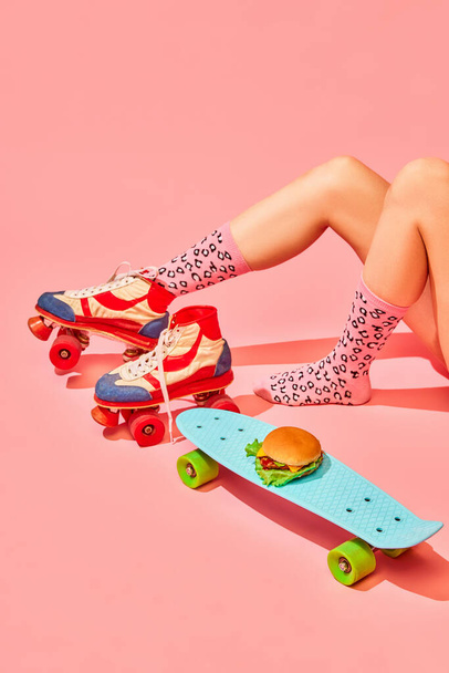 Female legs in vintage rollers and funny socks with delicious burger on skateboard against pink background. Delivery. Concept of pop art photography, creative vision, imagination. Minimal art - Foto, afbeelding