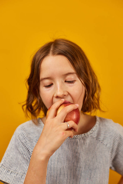 Pretty teen girl, child eat, bites a fresh red apple isolated on studio yellow background, healthy snack choices, wholesome food lifestyle concept, copy space - Foto, Bild