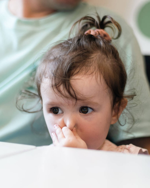 Une petite belle fille caucasienne pince le nez avec sa main et regarde attentivement sur le côté tout en étant assise dans les bras de son père à la table, vue latérale rapprochée. - Photo, image
