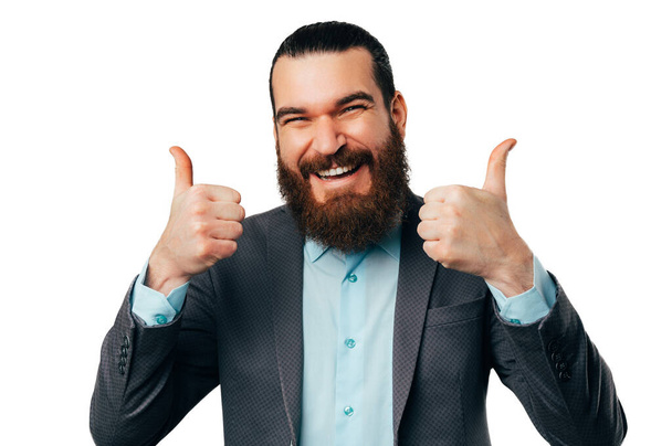 Close up portrait of an excited young bearded man holding two thumbs up over white background. - Zdjęcie, obraz