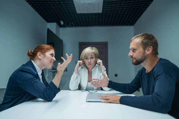 Блондинка, рыжая женщина и бородатый мужчина в костюмах в офисе. Деловые люди ругаются во время переговоров в конференц-зале - Фото, изображение